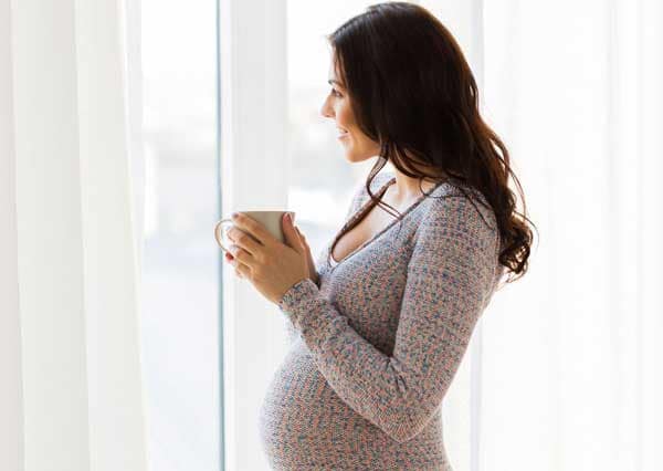 Schwangere-mit-Tasse-in-der-Hand-schaut-aus-dem-Fenster